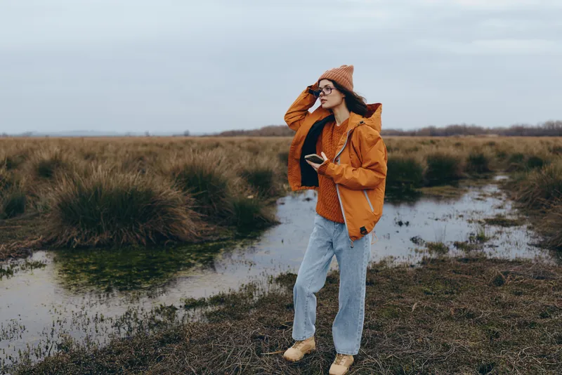 Woman lifestyle outdoors, in an autumn field, wearing a jacket, hat, sunglasses, and jeans, standing on a wetland path near water, cloudy sky, travel leisure mood, natural light