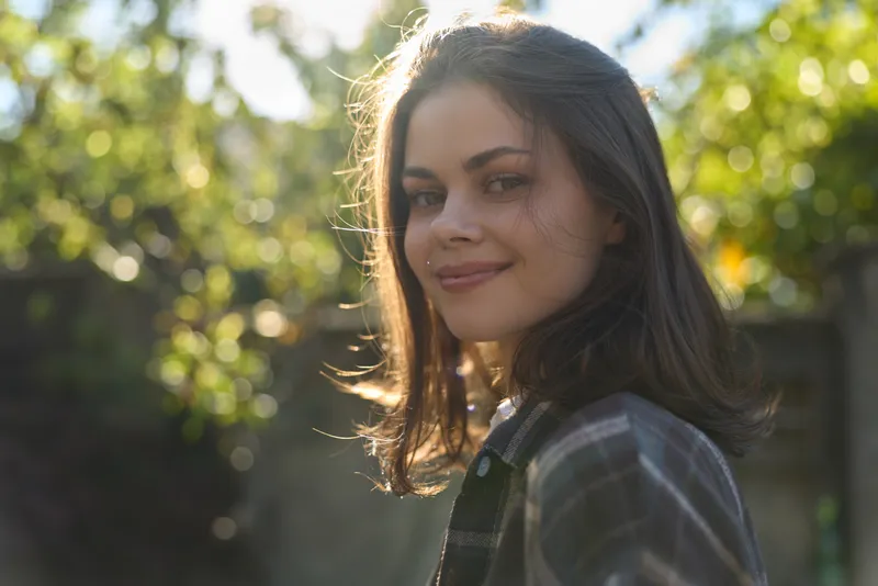 Smiling woman outdoors in a plaid shirt, natural garden backdrop, warm sunlight illuminating her face. Lifestyle portrait emphasizing relaxed, carefree mood and everyday beauty