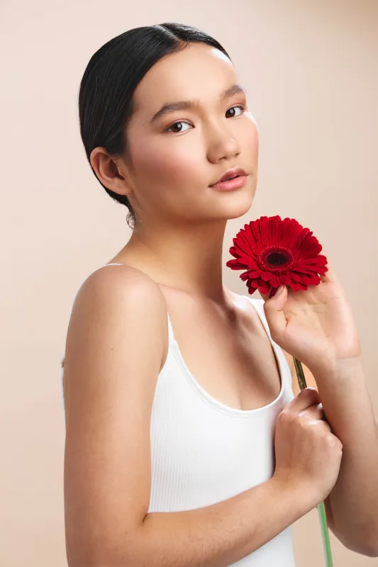 Asian woman in a minimalist studio holds a red flower near her face. She wears a white tank top, with a calm expression and soft lighting, creating a simple, elegant portrait.