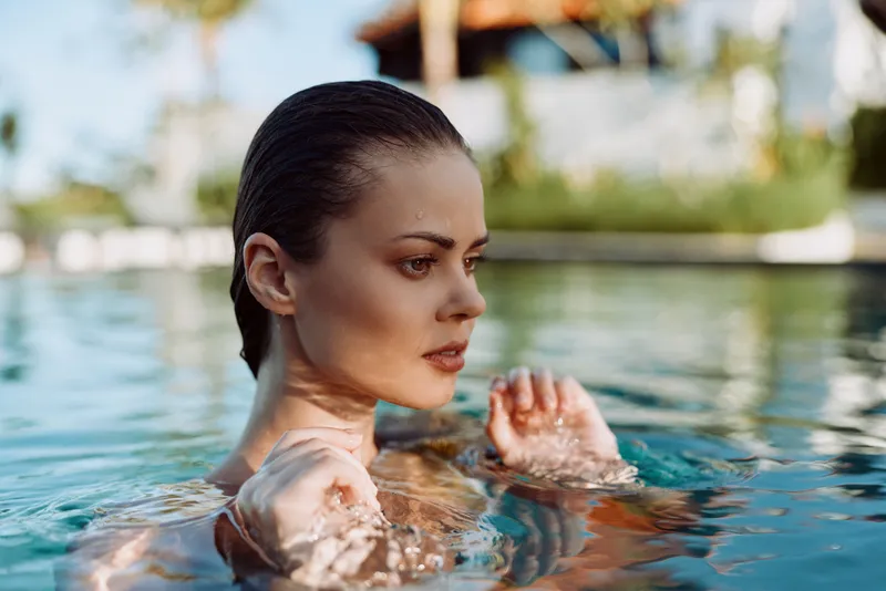 Woman swimming in a clear pool, head above water, focused face, sunlight on skin, relaxed summer mood, lifestyle and wellness moment, candid and serene scene by the water.