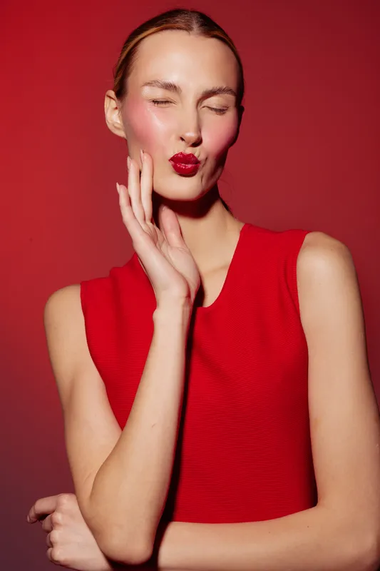 Young woman in red sleeveless top poses with eyes closed and puckered lips against red background. Concept of fashion, beauty, makeup, and confident female model portrait.