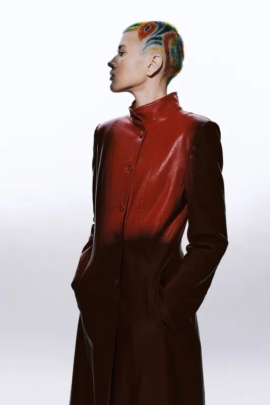 woman with colorful short hair wearing a long red leather coat, standing in profile on a bright white background, modern fashion style and strong confident pose