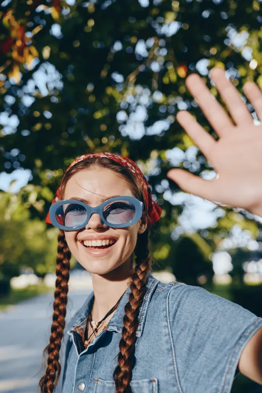 Lifestyle woman in modern boho-western denim street style wearing oversized sunglasses and red bandana, smiling outdoors with film color effect under sunny trees.