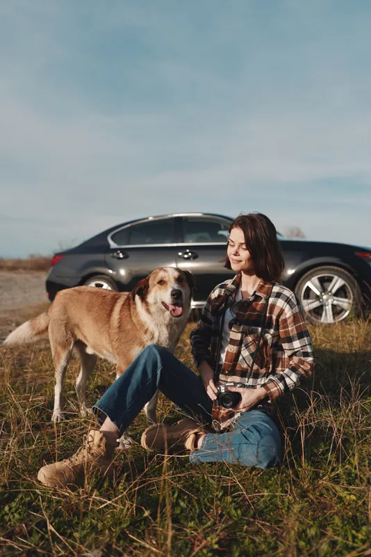 Woman sits in tall grass with a loyal dog beside a parked car in a sunny field, enjoying a quiet moment outdoors, casual fashion, and pet companionship, capturing a relaxed lifestyle scene away from