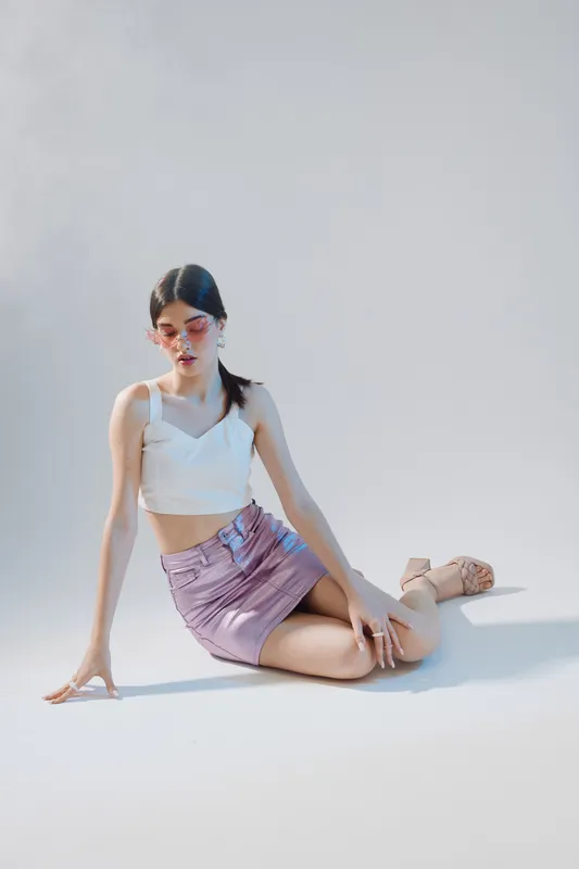 Young woman in a white top and pink skirt poses seated on the floor against a plain background. Concept of woman beauty fashion with soft natural lighting and minimalistic style.