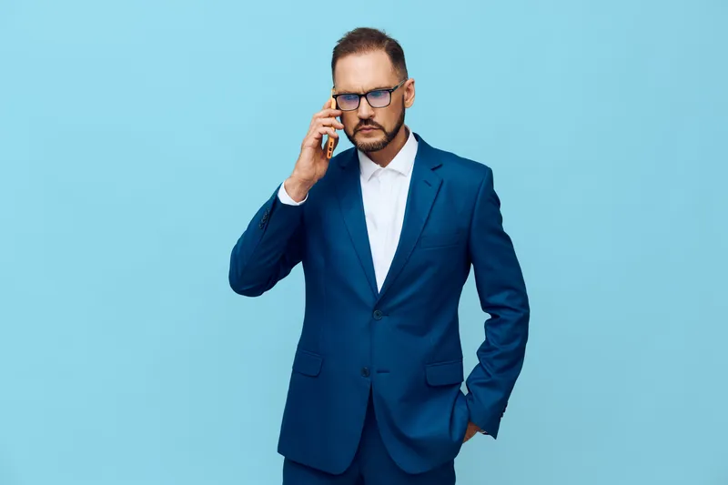 A professional business man stands against a solid blue background, wearing a tailored suit and glasses, while speaking on a mobile phone with a confident, focused pose.