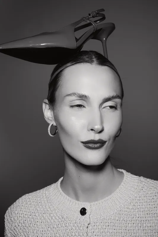 White Caucasian woman with pale skin and slicked hair balances a high heel shoe on her head. Studio portrait in black and white with minimalistic fashion concept.