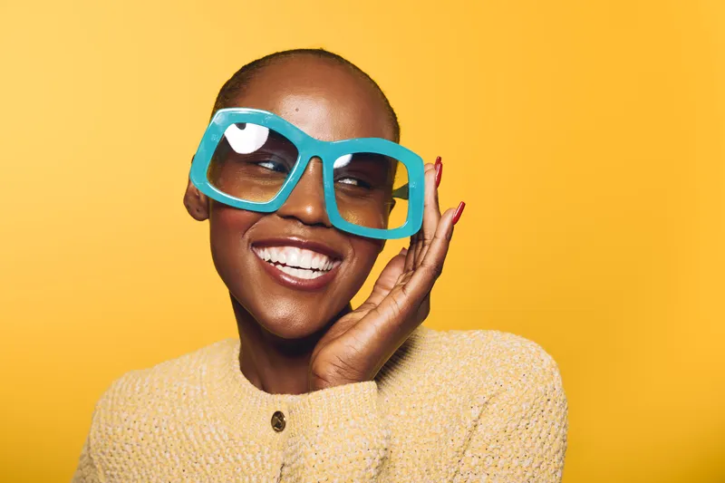 Happy African woman with dark skin wearing large blue sunglasses and yellow sweater smiles with eyes closed against bright yellow background. Concept for joy, fashion, and summer style.