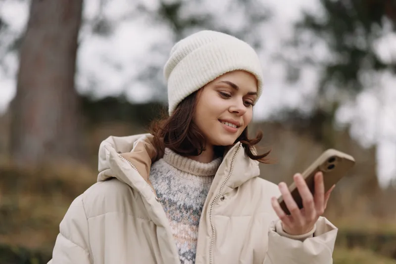 Woman in a winter coat and beanie checks her smartphone outdoors, smiling softly as she uses a mobile device in a chilly park setting with bare trees.