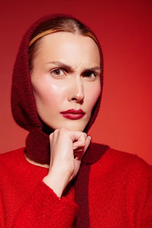 Portrait of a serious woman wearing a red knitted hood and sweater, looking thoughtfully with furrowed brows against a red background. Concept of contemplation and fashion.