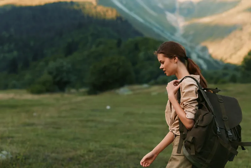 Young woman hiking outdoors with backpack walking through green meadow in mountains, casual outfit, natural light, focused expression, active lifestyle concept