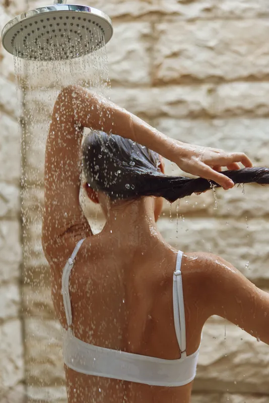 Woman taking a shower with water flowing from a modern showerhead, wet hair, and smooth skin under warm natural light.