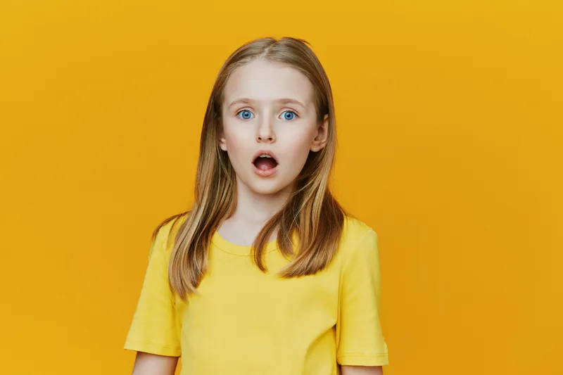 Surprised girl with blue eyes in a yellow shirt against a vibrant yellow background, expressing shock and wonder