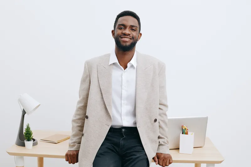 Confident young man with dark skin and beard wearing light beige blazer and white shirt, standing in modern office with white walls, desk, and laptop, looking at camera with friendly smile. People lif