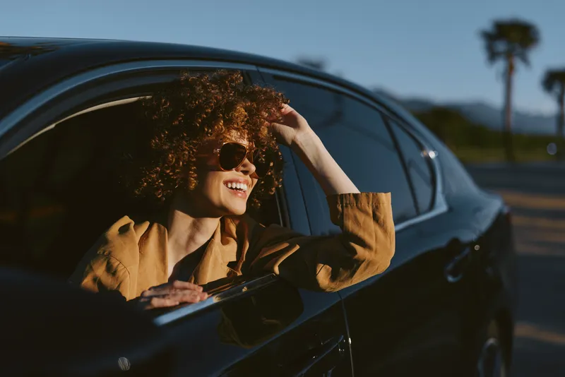 Smiling woman with curly hair in a car wearing sunglasses, enjoying a sunny drive. Retro film color enhances warm mood, casual lifestyle, and sense of freedom.