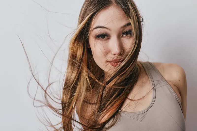 Portrait of young woman with long hair blowing in wind, wearing casual sleeveless top. Studio background with natural makeup and confident expression closeup on face and hair.