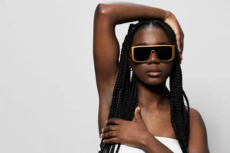Stylish black woman with braided hair wearing oversized sunglasses, showcasing modern fashion and confidence against a minimalist background