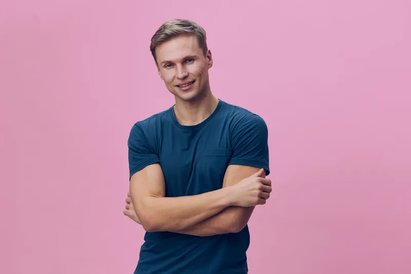 Cheerful smiling young man with short blond hair wearing casual blue t-shirt posing with arms crossed against pink background. Studio portrait, lifestyle concept, positive emotions, youthful energy, v