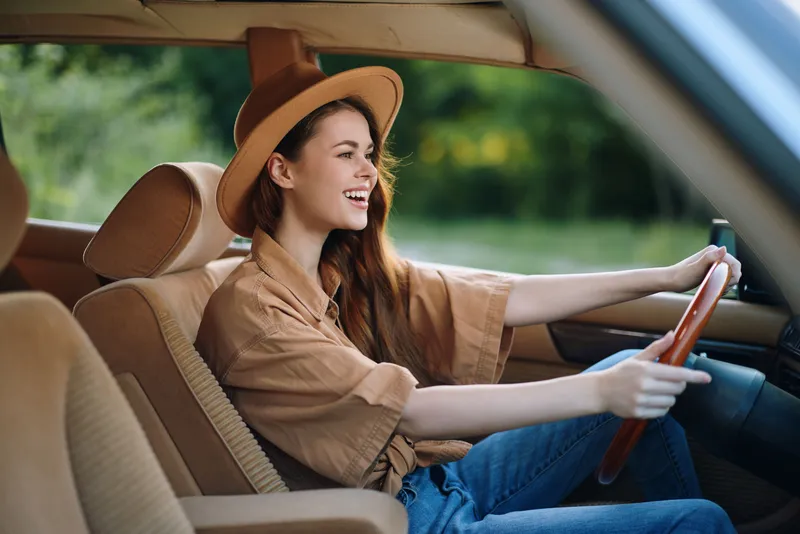 Happy young woman driving a vintage car, wearing a stylish hat and casual outfit, in a lush green background showcasing freedom and summer vibes.