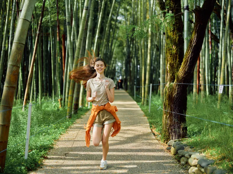 Woman running on a gravel path through a bamboo grove smiling in casual summer clothes with an orange jacket tied at waist, active travel and joyful nature trail scenery.
