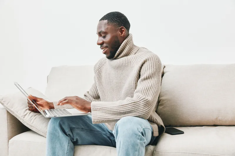 Young African man wearing a cozy sweater, joyfully working on a laptop on a sofa in a minimalist living room, emphasizing productivity and comfort