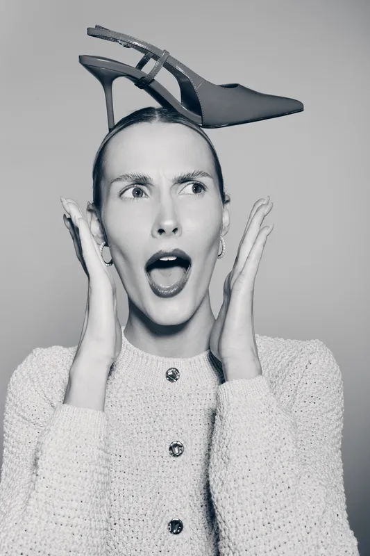 Surprised white woman with pale skin balances a high heel shoe on her head, wearing knitted sweater, studio portrait.