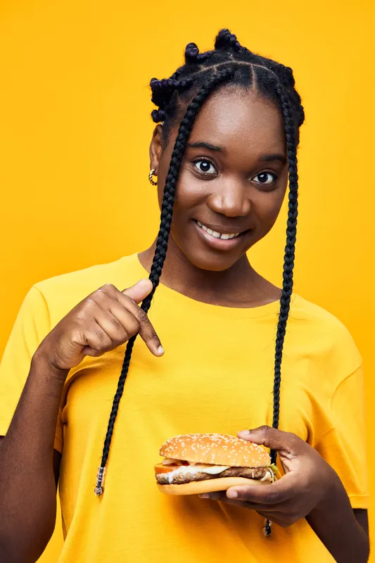 Smiling young African girl pointing at a delicious burger, wearing a bright yellow shirt against a vibrant yellow background, conveying a positive and cheerful vibe
