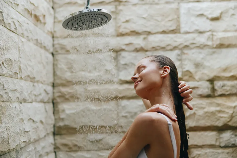 woman enjoying shower water in stone modern bathroom, relaxed mood, wet hair, casual underwear, natural light, self-care moment