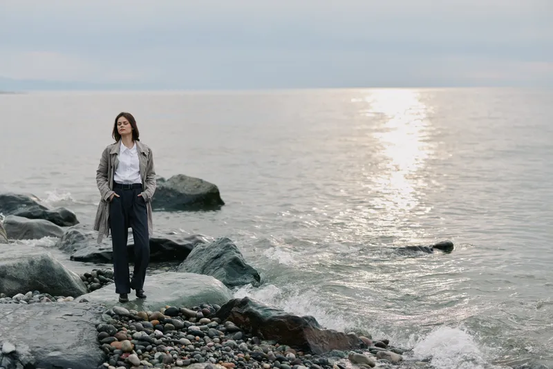 Young woman stands on rocky beach wearing casual outfit, gazing at calm sea under cloudy sky. Concept of solitude, nature, relaxation, and peaceful outdoor moment.