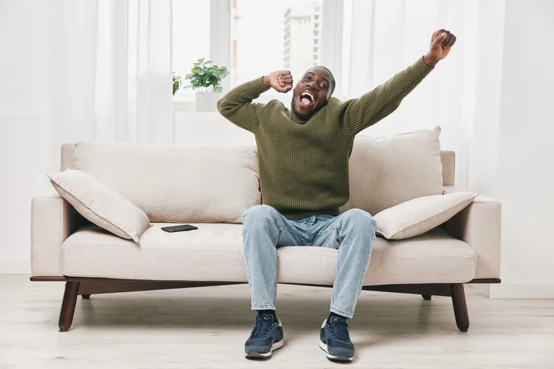 Cheerful African man stretching on a cozy sofa in a bright living room, wearing a green sweater and blue jeans, expressing joy and relaxation