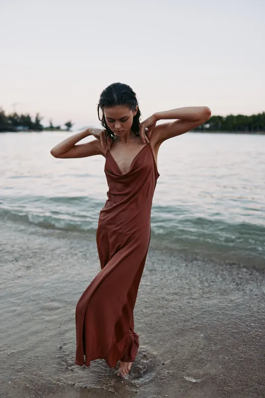 A woman in a rust brown slip dress stands in shallow beach water, sleeves lifted as waves brush her feet, while a calm horizon and soft light frame the scene.