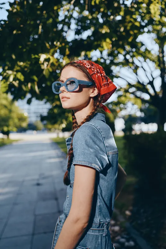 Modern boho-western denim street style woman wearing blue sunglasses and red bandana, lifestyle portrait with film color effect in natural urban background lighting.