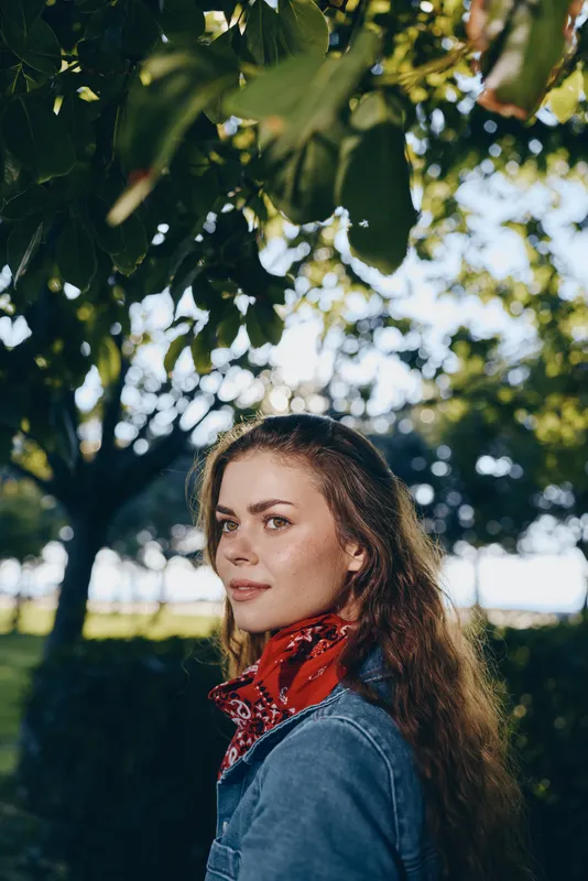 Lifestyle woman in modern boho-western denim street style with a red bandana, natural light, and film color effect capturing relaxed outdoor vibes under leafy trees.