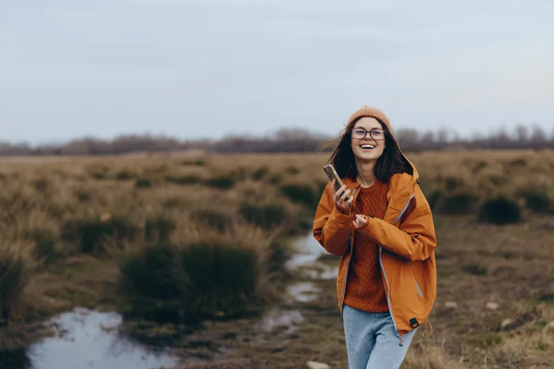 Smiling woman lifestyle outdoors in a rural field, wearing orange jacket and jeans, standing on a path, soft daylight, nature background, casual travel, wide landscape view, with glasses