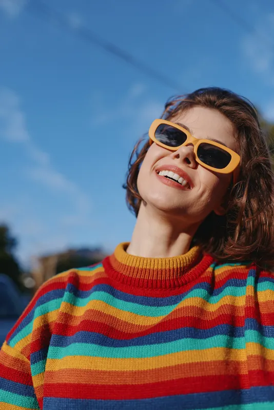 Woman smiling in a colorful striped sweater and yellow sunglasses, outdoor sunny portrait of a mid adult casual model leaning back with joyful expression for travel lifestyle fashion use.
