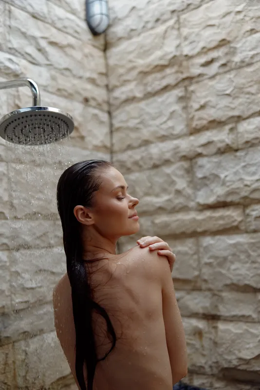 Relaxed woman enjoying outdoor shower with wet hair and eyes closed, standing against stone wall with gentle water flow, natural and serene atmosphere.