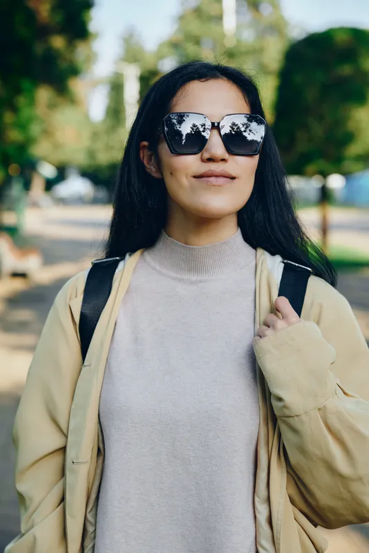 Sunglasses, woman, outdoor park fashion in a beige coat and light sweater, carrying a backpack, relaxed pose, confident mood, street style in natural daylight and greenery