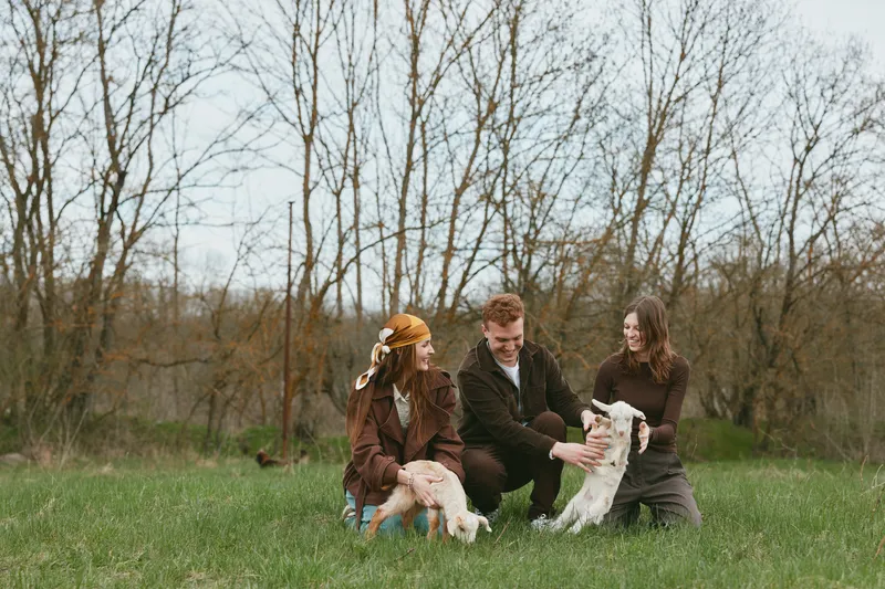 A cheerful family enjoys a sunny outdoor moment with a goat and kid in a grassy field, showing farm life, affection, and playful bonding in a natural countryside setting.