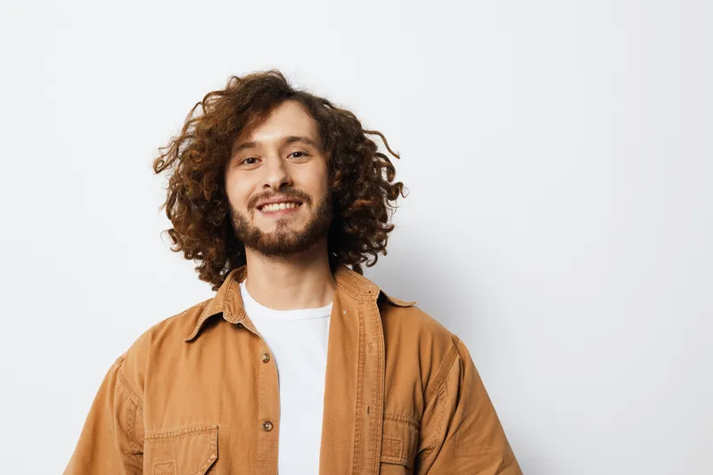 Young man with curly hair smiling in a casual outfit, displaying happiness against a minimalist white background, perfect for lifestyle and personal branding themes
