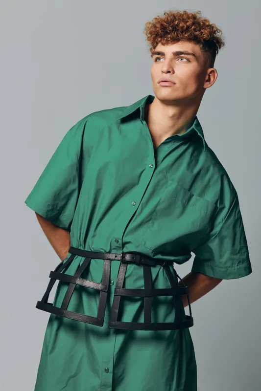 Young man with curly hair wearing oversized green shirt and black cage belt posing against gray background. Fashion model with serious expression and modern style in studio portrait.