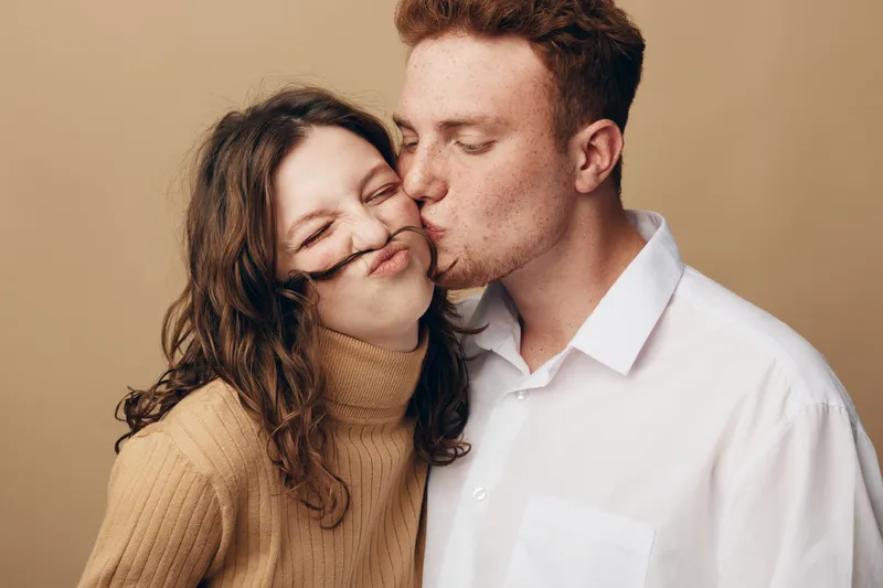 Young couple in casual clothes share a tender moment as the man kisses the woman on the cheek, creating a playful and loving atmosphere on a neutral background.