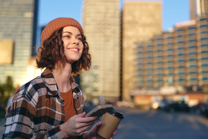 Woman with coffee and phone in city, smiling portrait at golden hour; urban morning scene capturing authenticity, candid lifestyle, mindful living and emotional storytelling in warm sunlight.