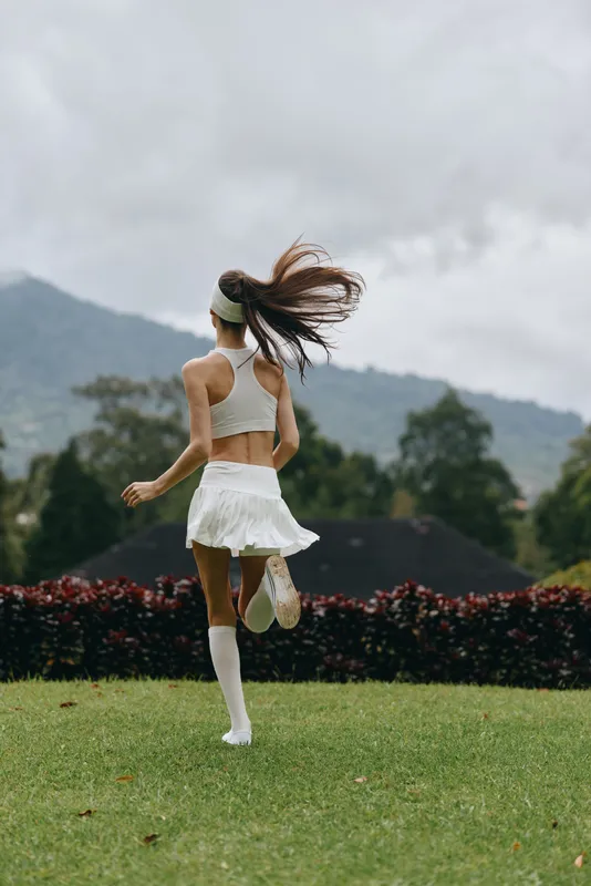 Young athletic woman in a white sports outfit running outdoors with long hair flying, showcasing freedom and joy in a lush green landscape