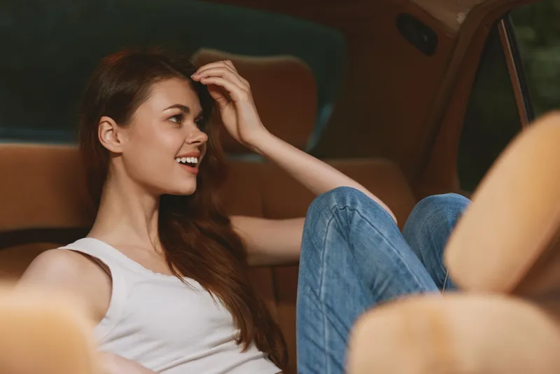 Smiling young woman relaxing in a car's backseat, wearing casual clothes, conveying joy and comfort amidst a warm brown interior.