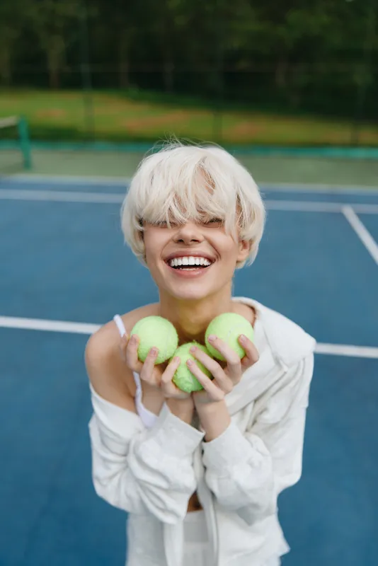Young woman with a short blonde hairstyle joyfully holding green apples on a tennis court, exuding a vibrant and healthy lifestyle