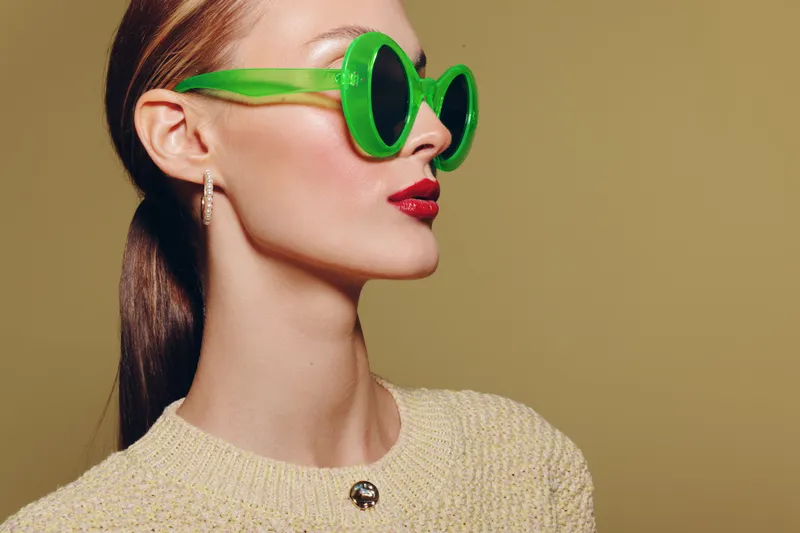 Stylish woman with green sunglasses and red lipstick poses in beige sweater against plain background. Fashion portrait with bold accessories and modern makeup look.