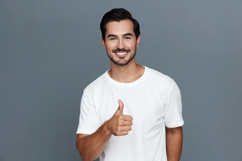 Smiling man in white t-shirt showing thumb up, looking at the camera against solid grey background. Positive vibes and cheerful lifestyle concept.