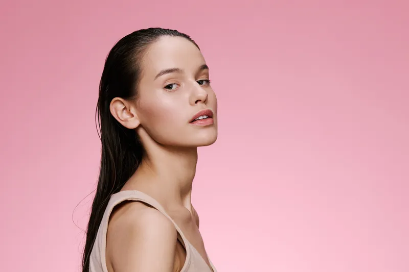 Young woman with wet hair, natural makeup, and neutral clothing against a soft pink background, embodying beauty and confidence in a fresh and vibrant concept