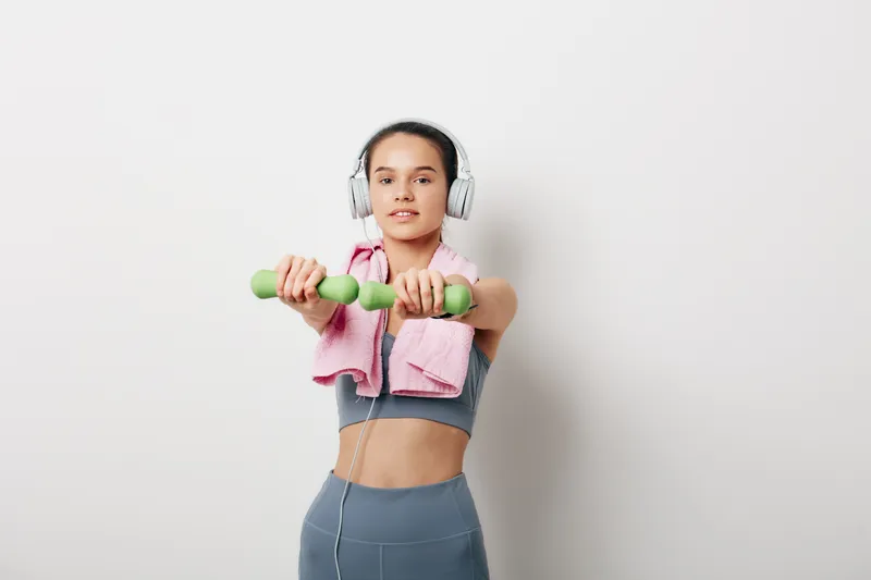 Young woman fitness portrait performing a front raise with green dumbbells, wearing headphones and a pink towel over shoulders, gray sports bra and leggings against a clean white background.