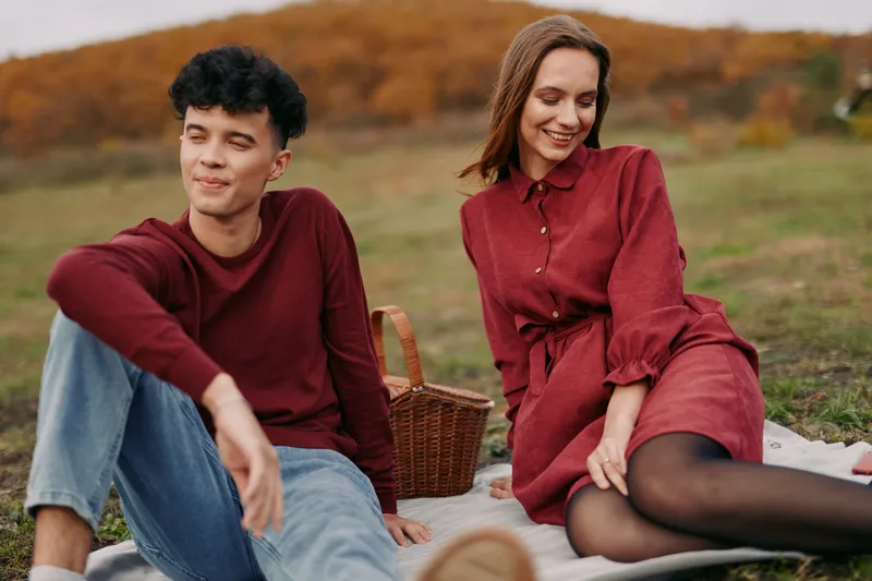 A candid outdoor portrait of a young couple on a picnic in an autumn field, conveying authenticity and credibility through genuine smiles, relaxed poses, unposed interaction, and warm tones.